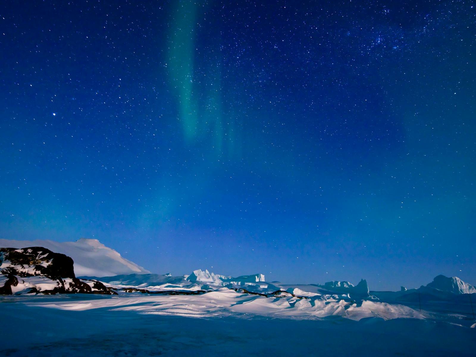 Aurora Australis glows in the night sky above a snowy, mountainous landscape.
