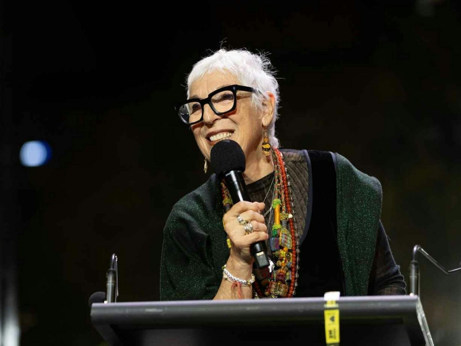 A woman with short grey hair and glasses holding a microphone on stage
