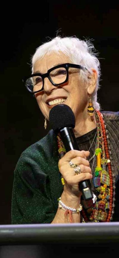 A woman with short grey hair and glasses holding a microphone on stage