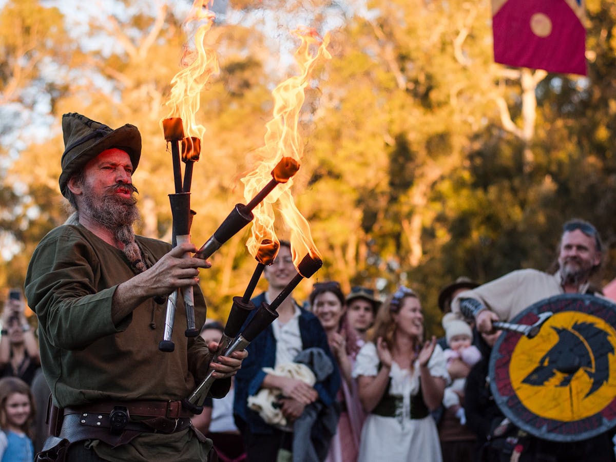 Performer with flaming juggling sticks grimaces. A viking in the background is clanging an axe.