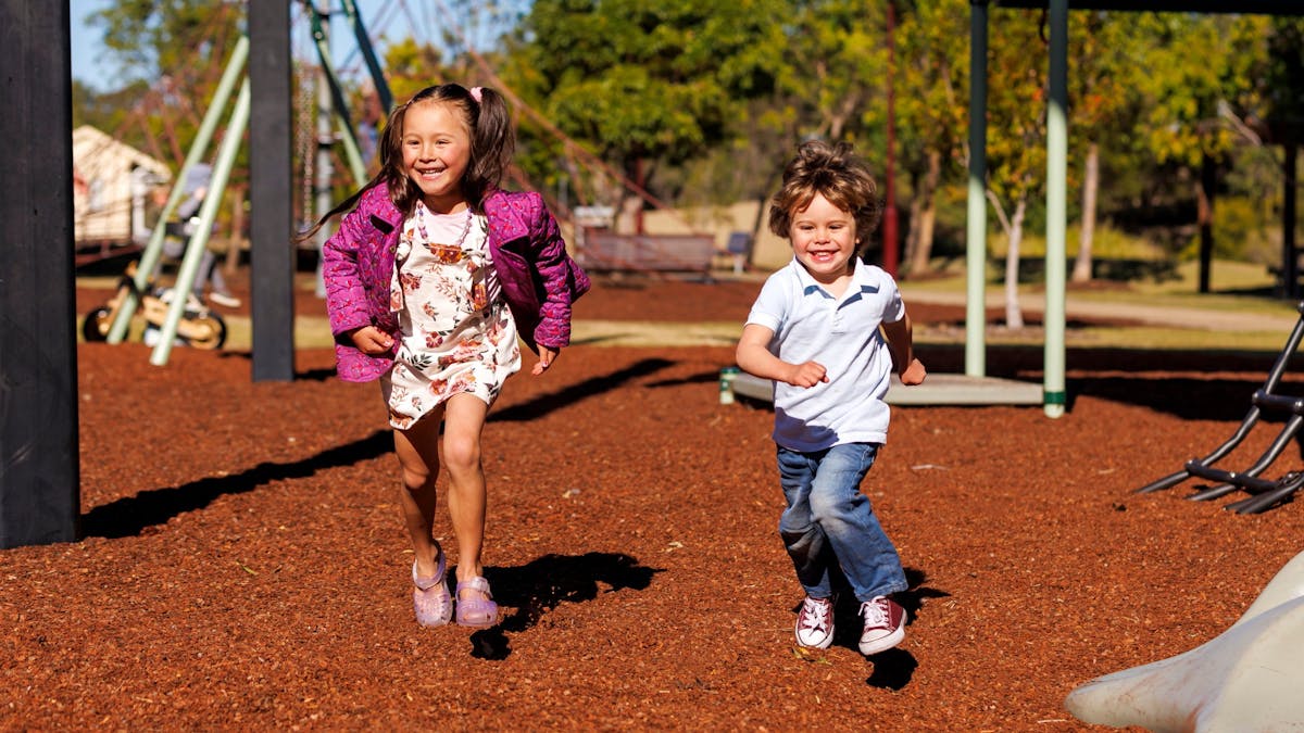 Girl and boy playing in the park with playground in the background at Pipeliner Park