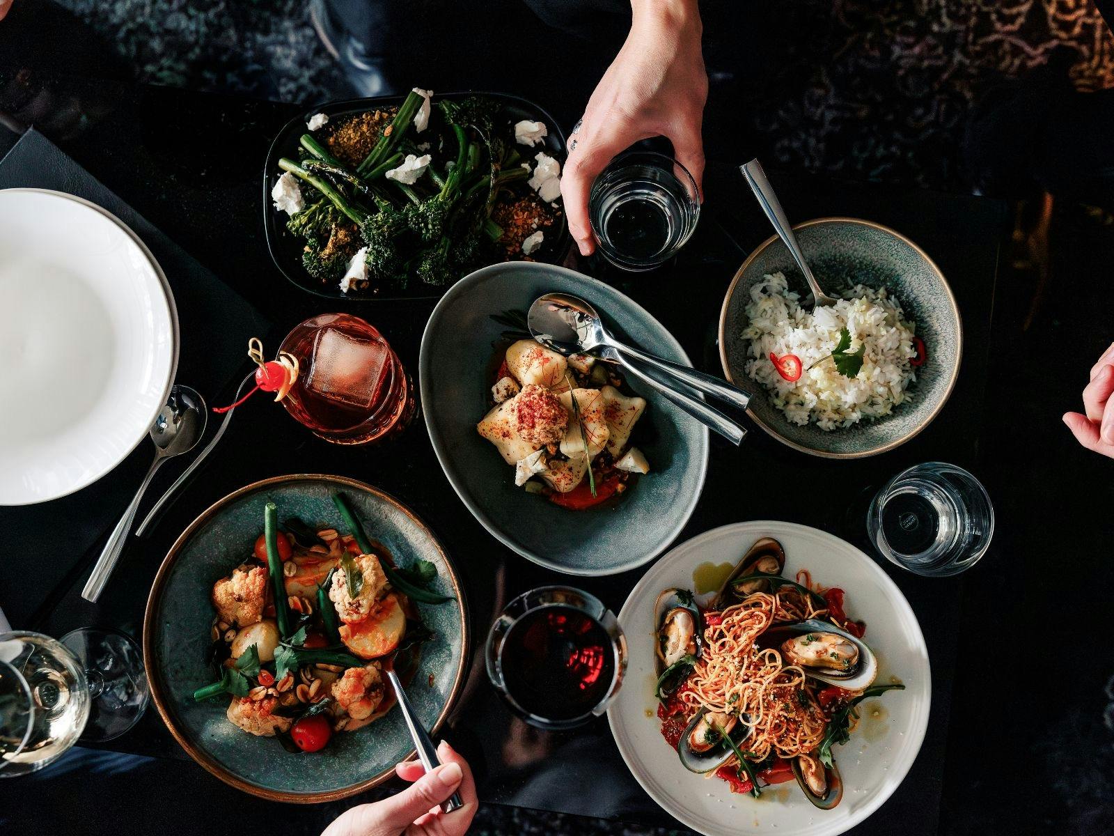 Multiple dishes of rice, pasta, curry, sides with hands eating, glasses cheering