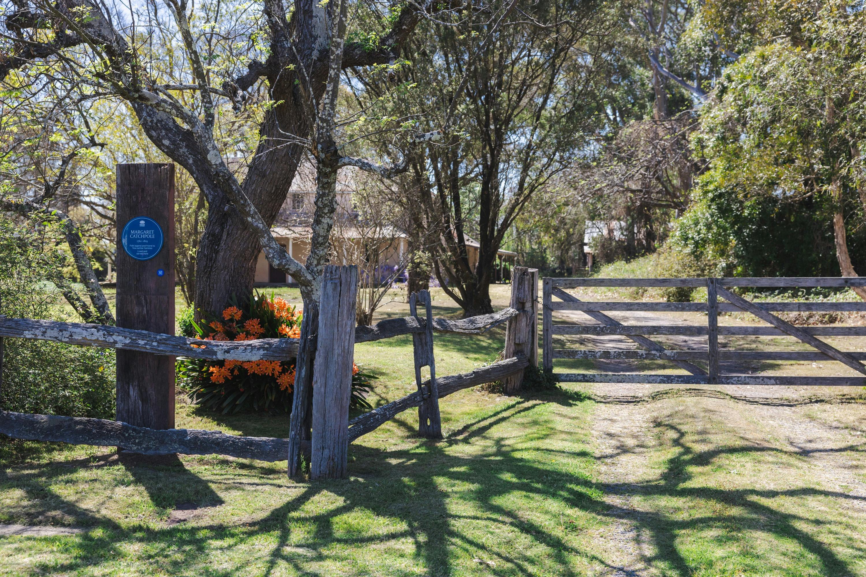 Blue plaque on post, garden behind timber fence, driveway to two-storey brick house