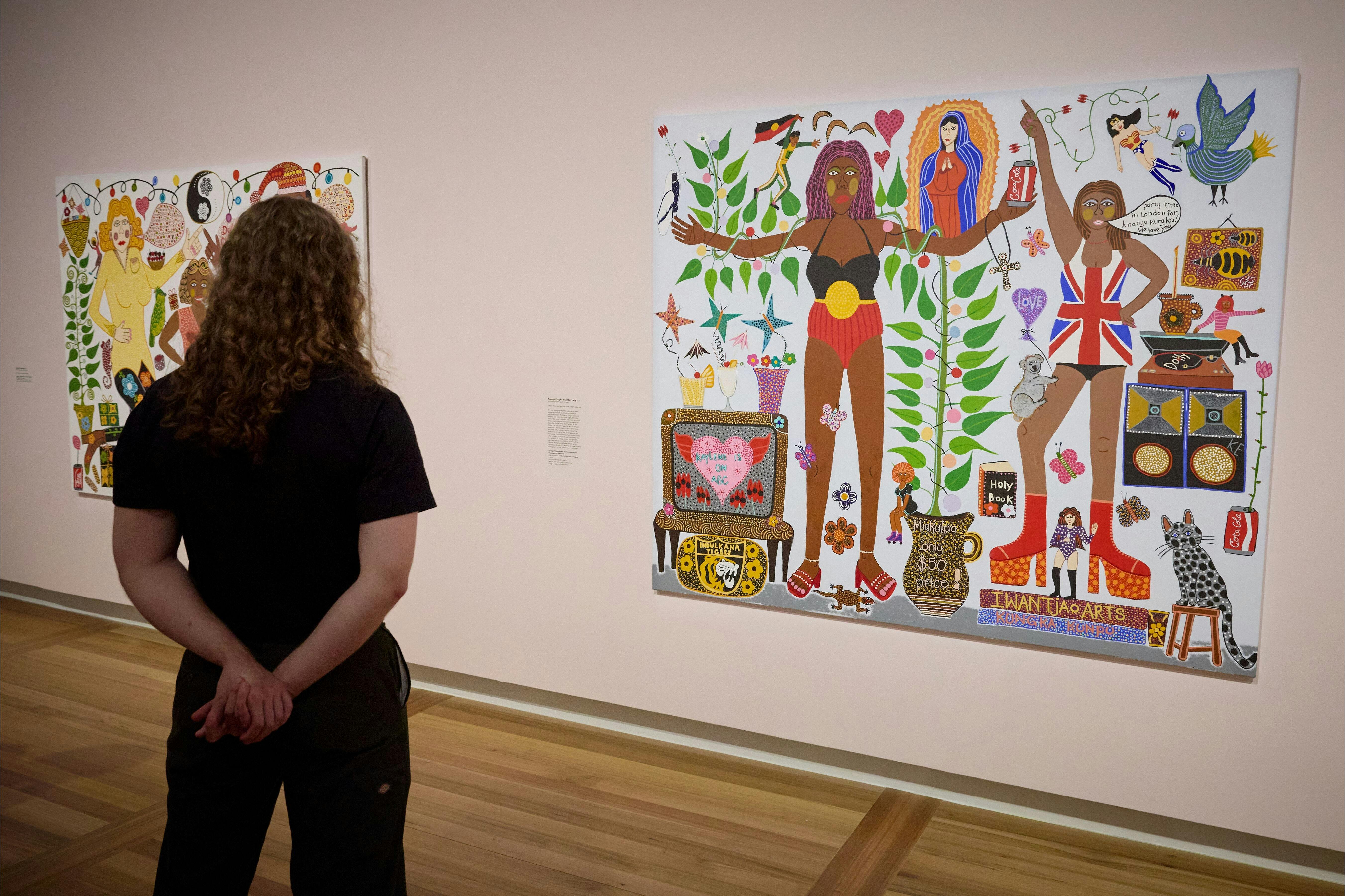 A woman looks at a colourful, eclectic portrait by Kaylene Whiskey that hangs on a pale pink wall.