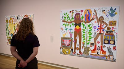 A woman looks at a colourful, eclectic portrait by Kaylene Whiskey that hangs on a pale pink wall.