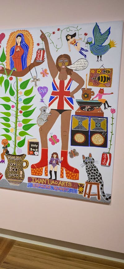 A woman looks at a colourful, eclectic portrait by Kaylene Whiskey that hangs on a pale pink wall.