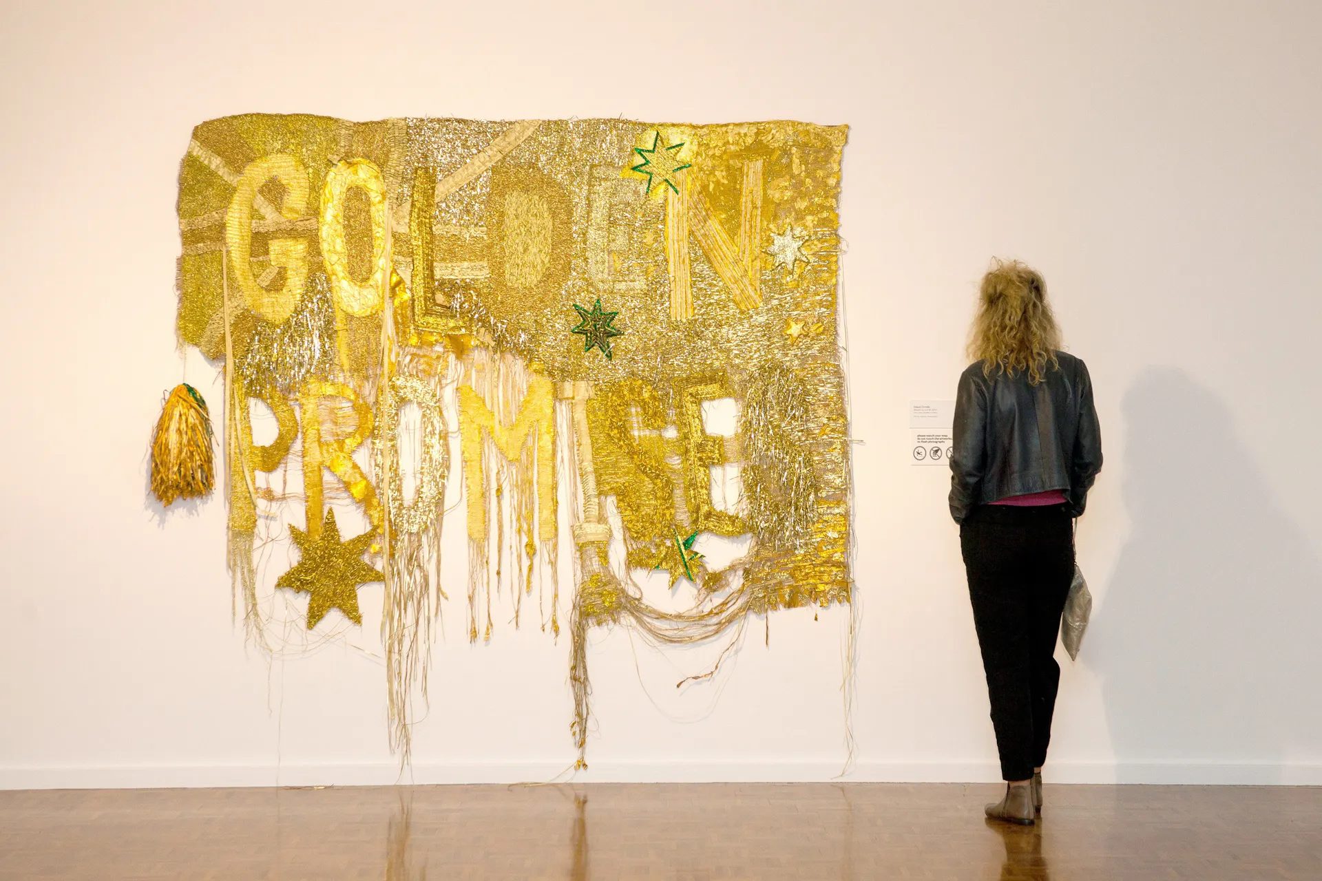 A woman dressed in black views at a large gold tapestry baring the words Golden Promise