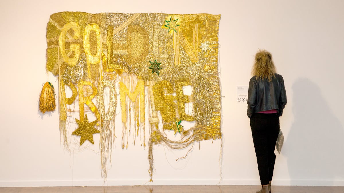 A woman dressed in black views at a large gold tapestry baring the words Golden Promise