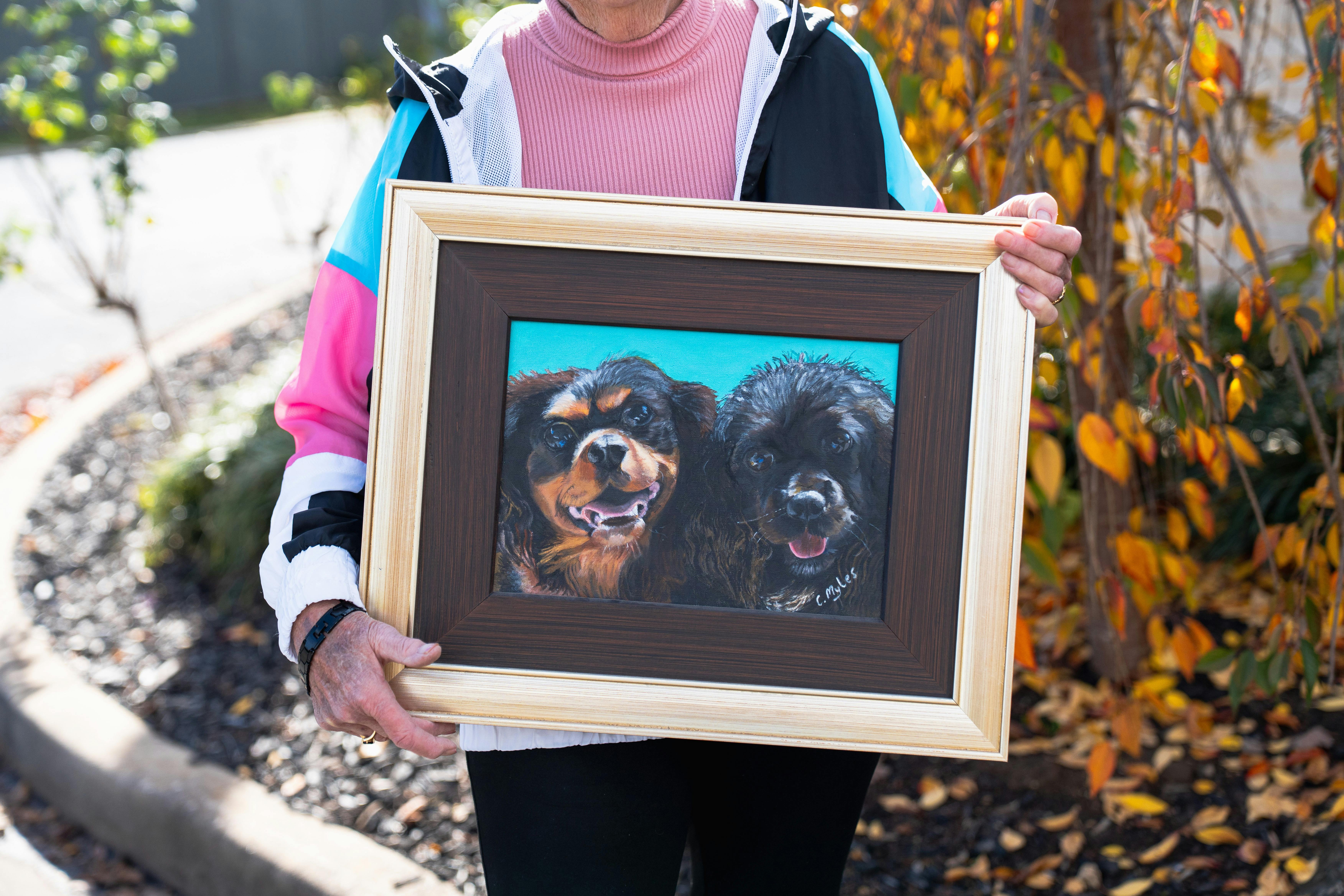 Older artist holding a framed painting of two dogs outdoors at Barossa Village.