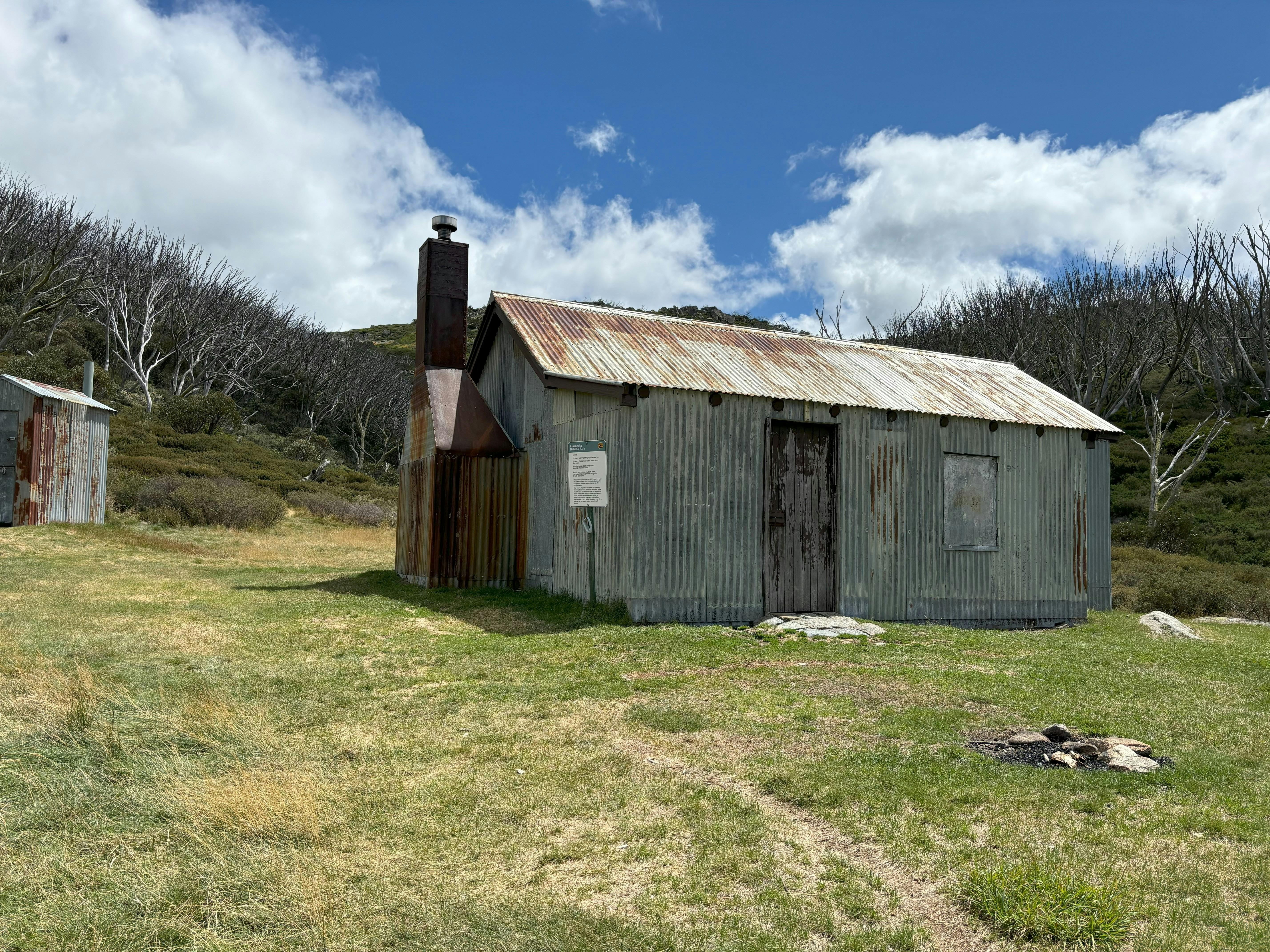 A small corrugated iron mountain hut and out side toilet in front of a stand of snow gums.