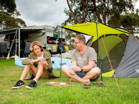Dad and son playing cards on lovely green grass  in front of their tent, caravans in the background