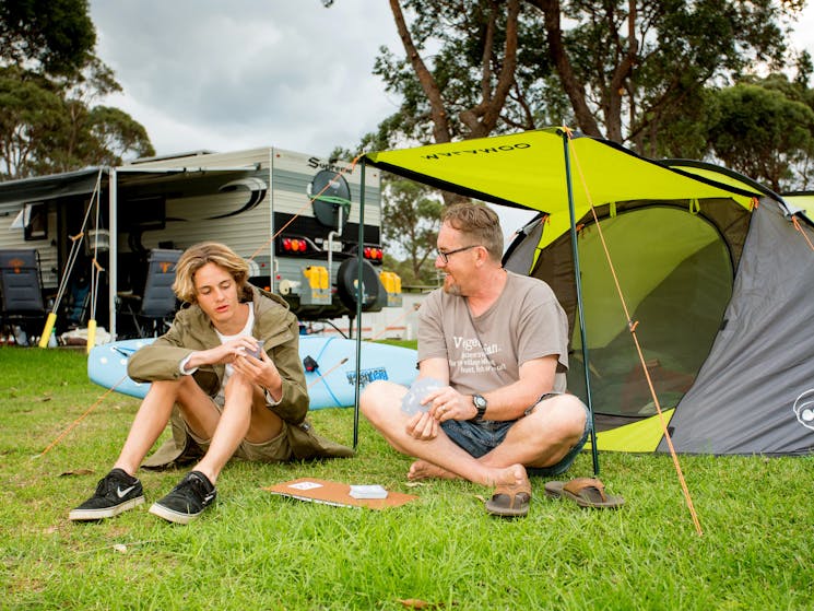 Dad and son playing cards on lovely green grass  in front of their tent, caravans in the background