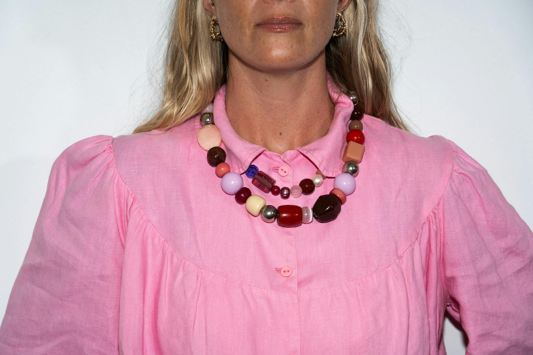 A blonde woman with a pink shirt wears 2 beaded necklaces