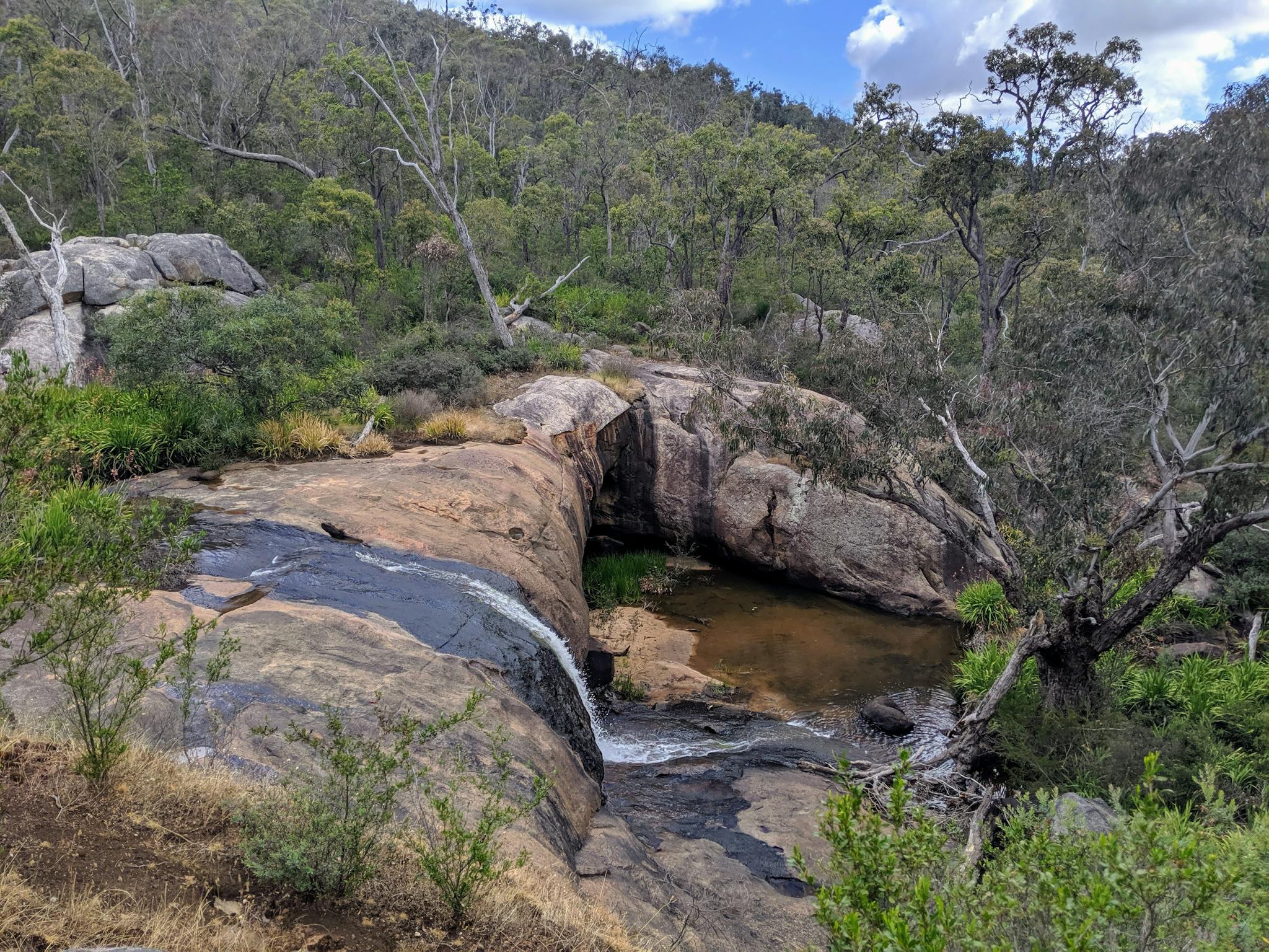Waterfall over large smooth boulders with a small hill covered with trees in the background.