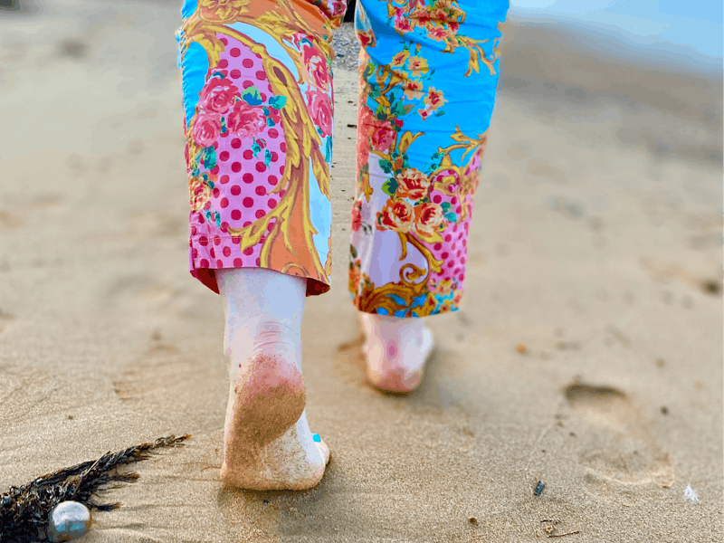 Bare feet imprinting the sand on Penguin Beach, by woman wearing colourful trousers.