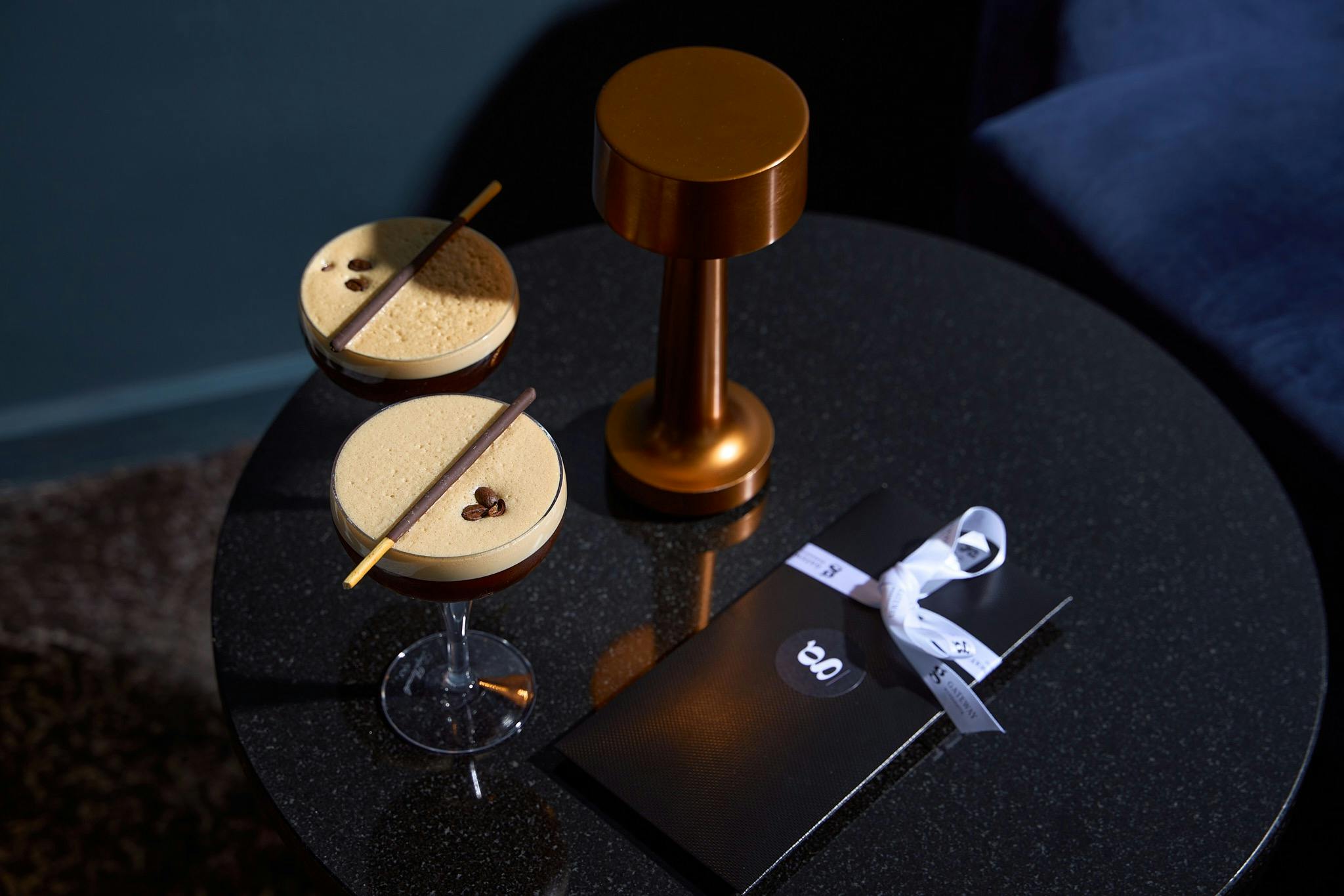 Two espresso martini cocktails, next to a candle, gift voucher and blue velvet chair