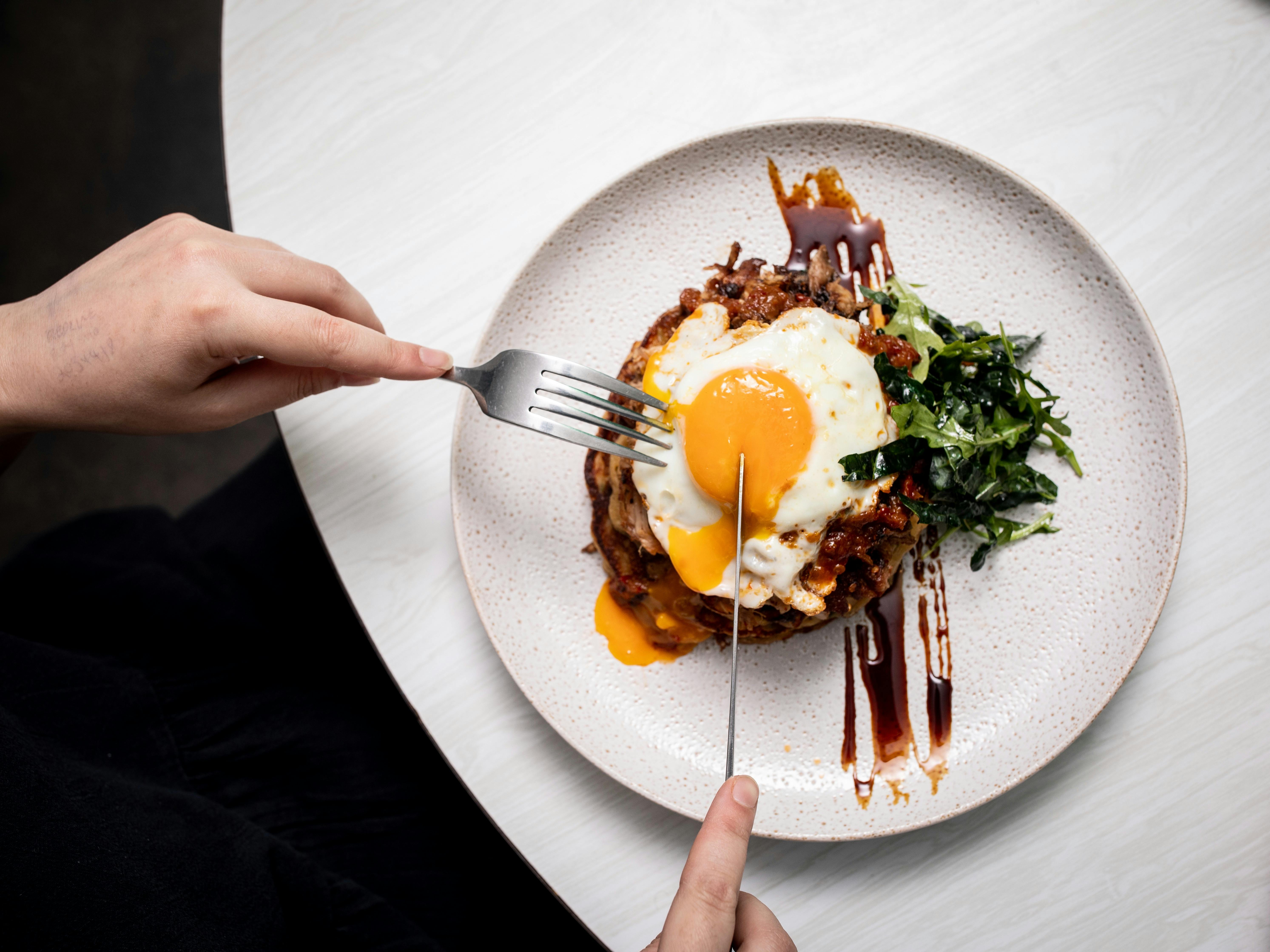 Pork Sword Breakfast - two hands holding cutlery, cutting into a fried egg  breakfast on a plate