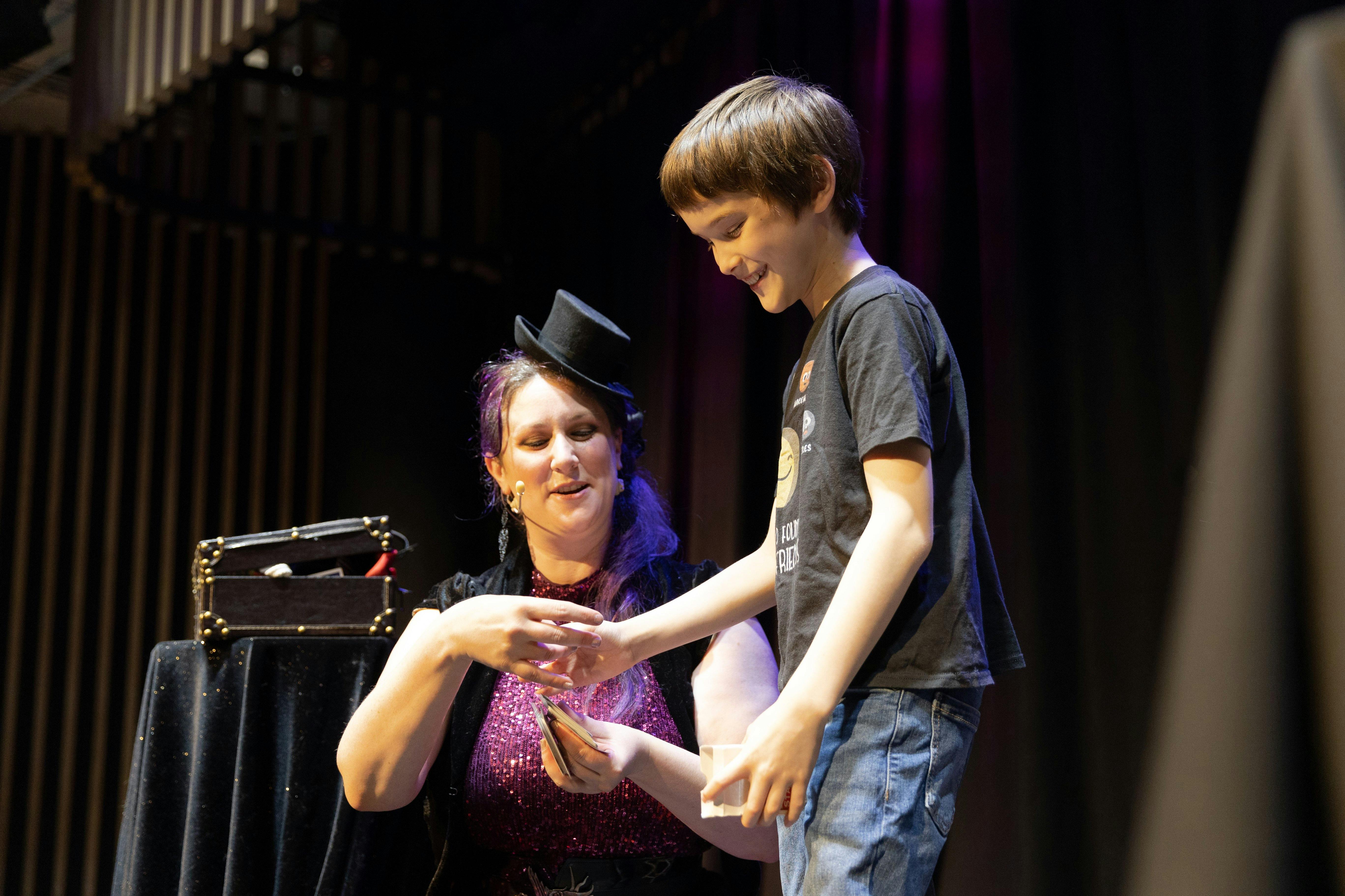 One magician bent down, assisting a young child with a deck of cards. Black curtain in background.