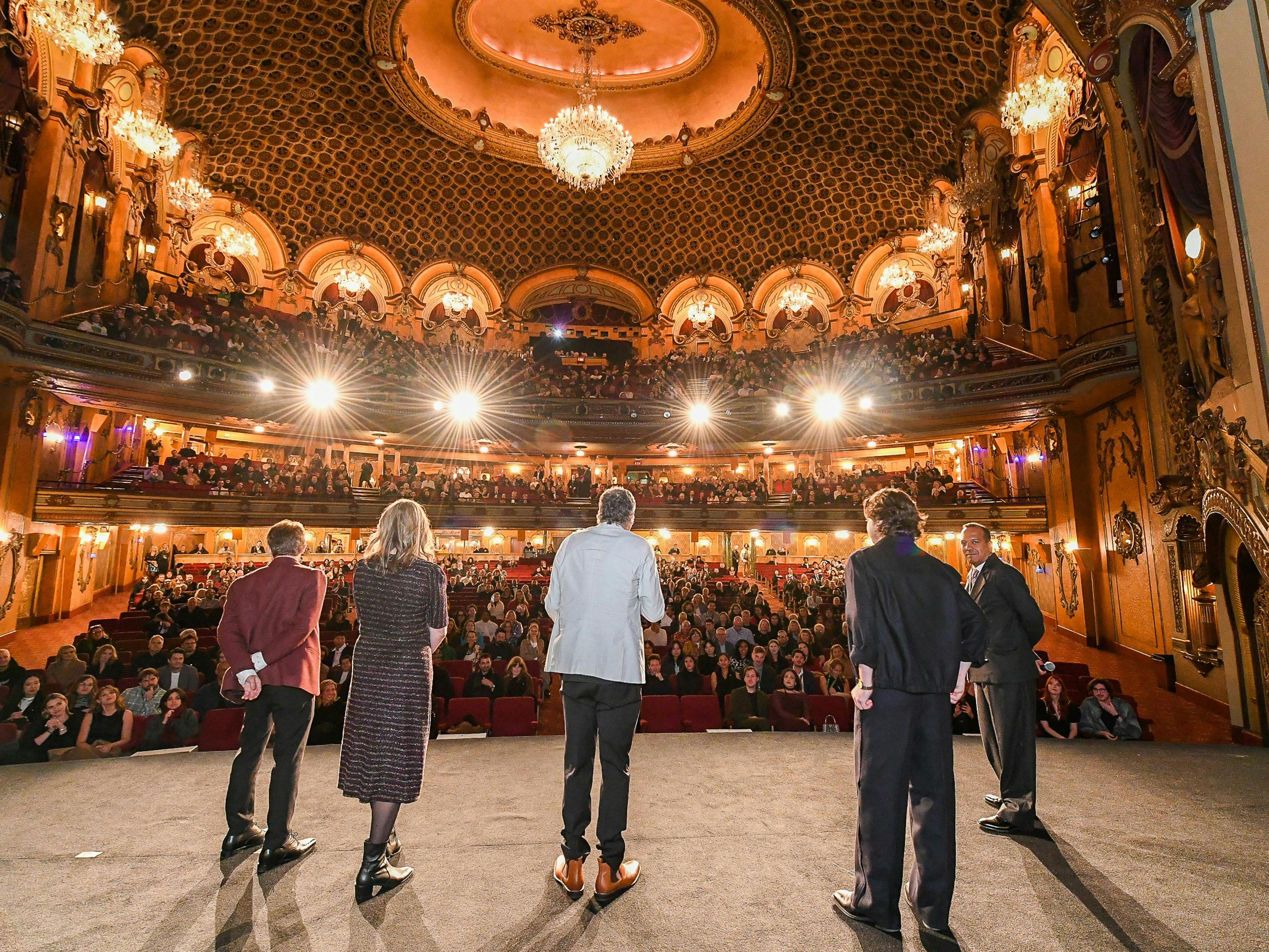 Five people look out from a stage to a huge crowd in a glamorous theatre.