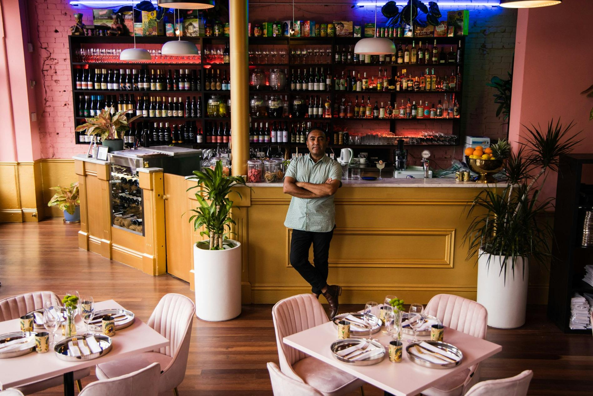The owner Jessi standing up against the bar, with table in the foreground & a stocked bar behind