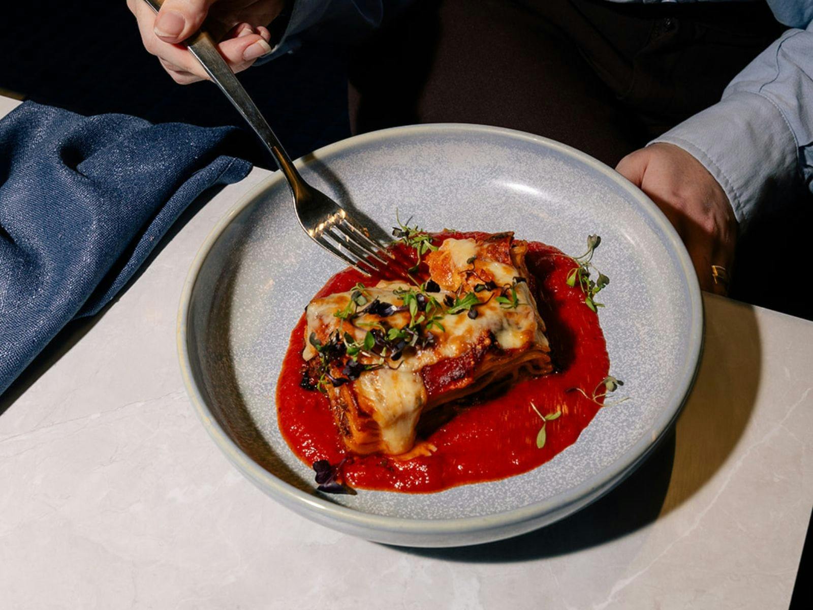 Fork going into a slice of lasagne in bowl on a table