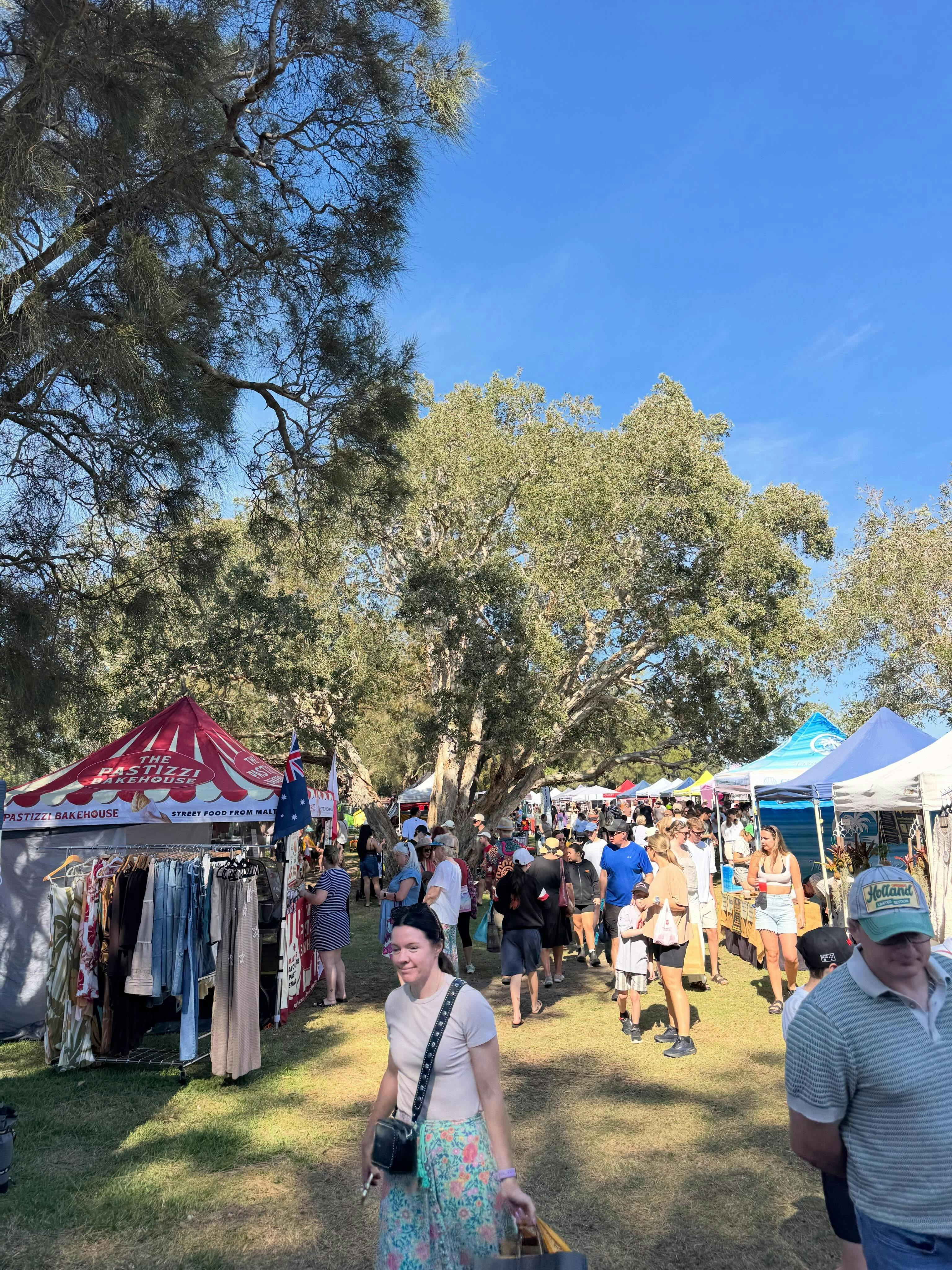 Visitors browsing colourful market gazebos beneath tall trees on a sunny day.