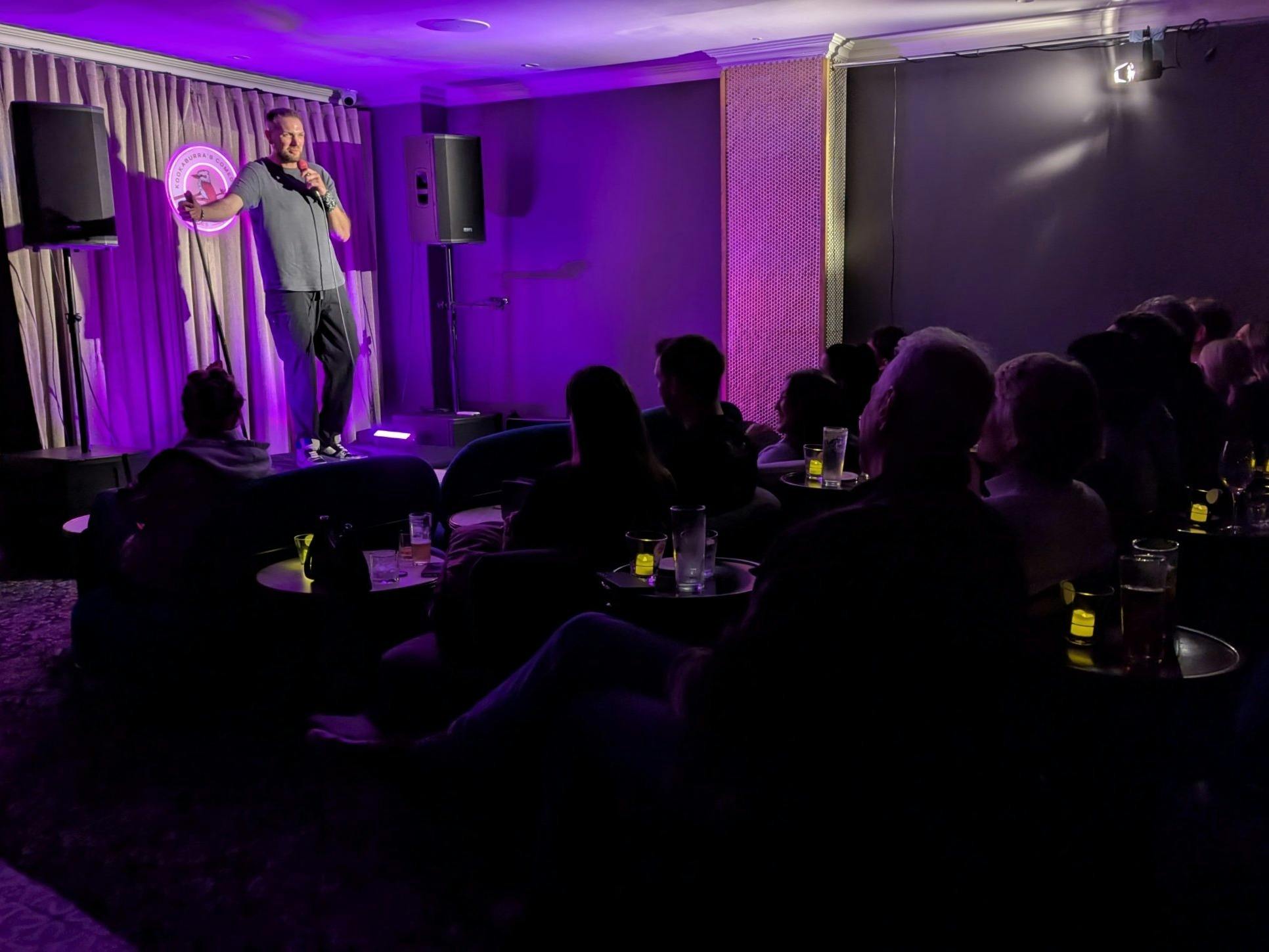 A stand-up comedian on a small stage with a microphone, facing an audience seated at low tables