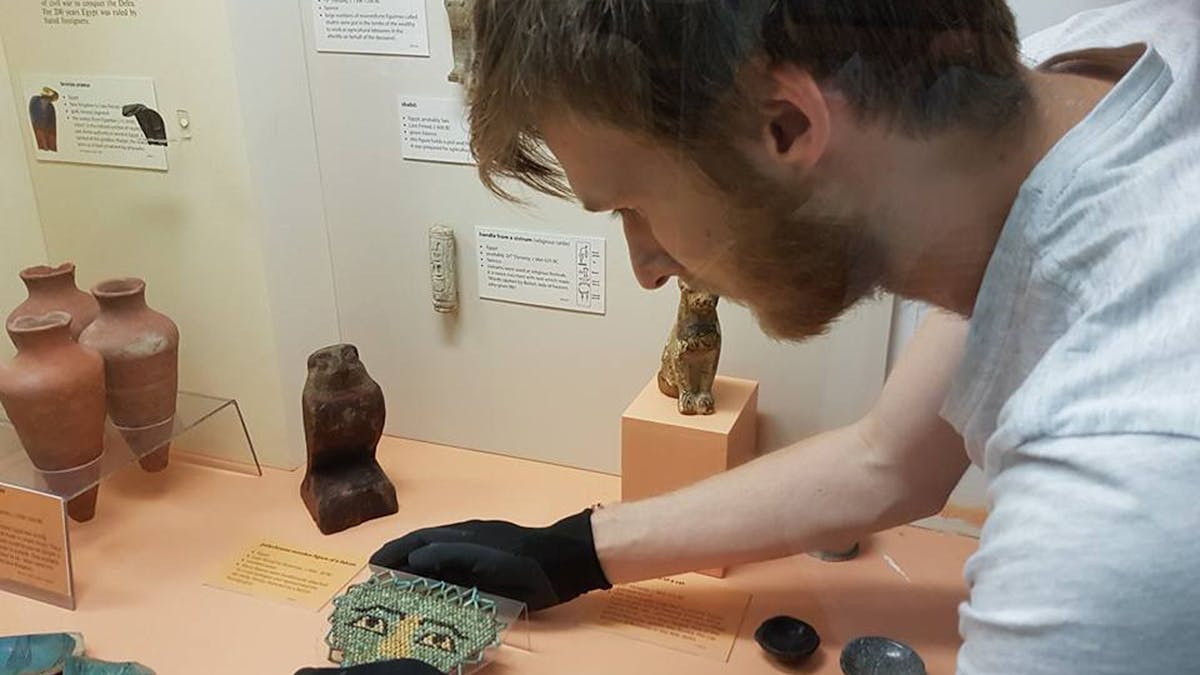 A view into the Egyptian Display Case showing staff with gloves arranging artefacts and  labels .