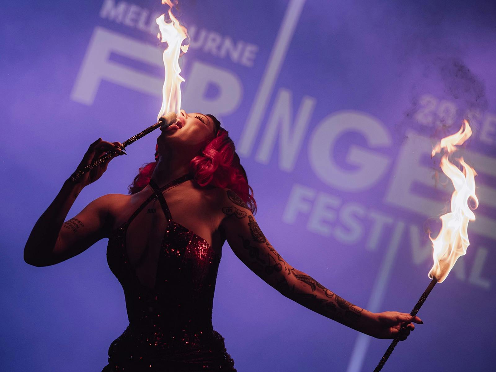 A performer in a dark sequined outfit holds two flaming batons, one at their mouth.