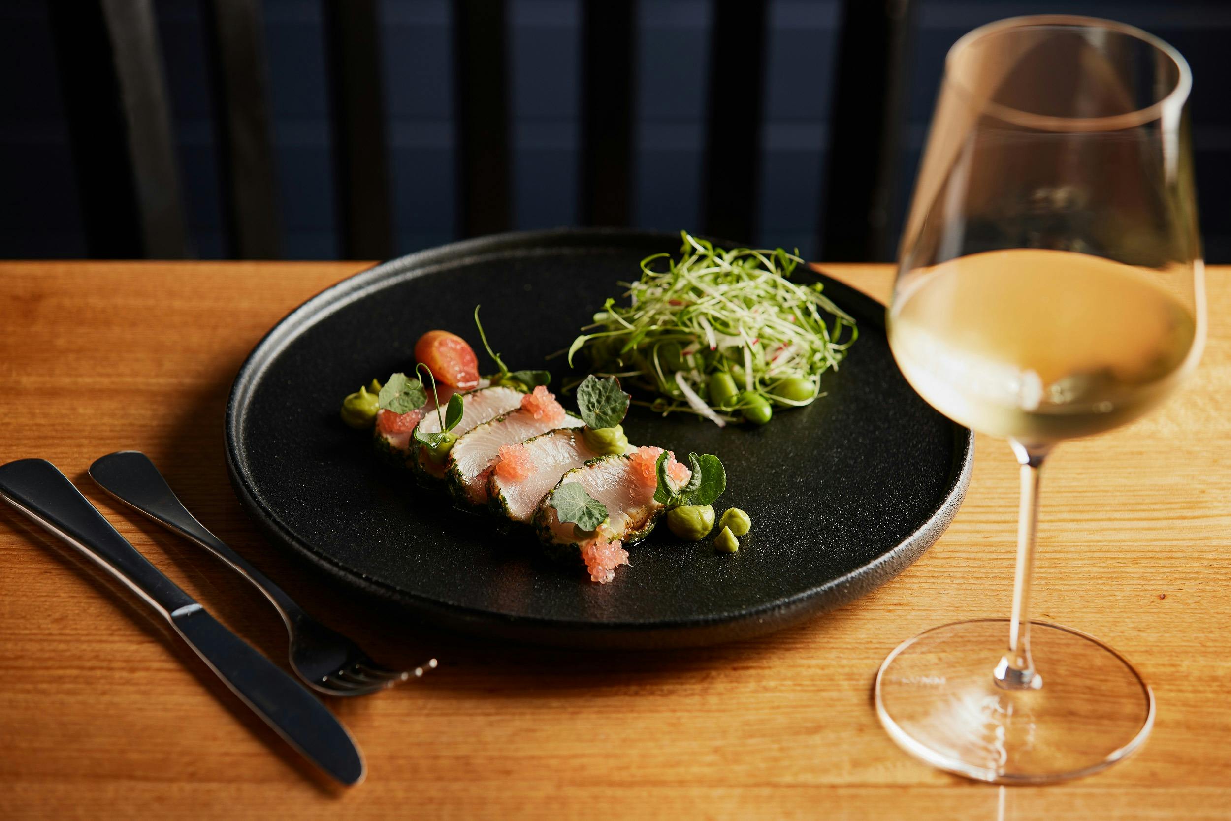 Australia Kingfish, thinly slice, presented on a black plate with a small salad and glass of wine.