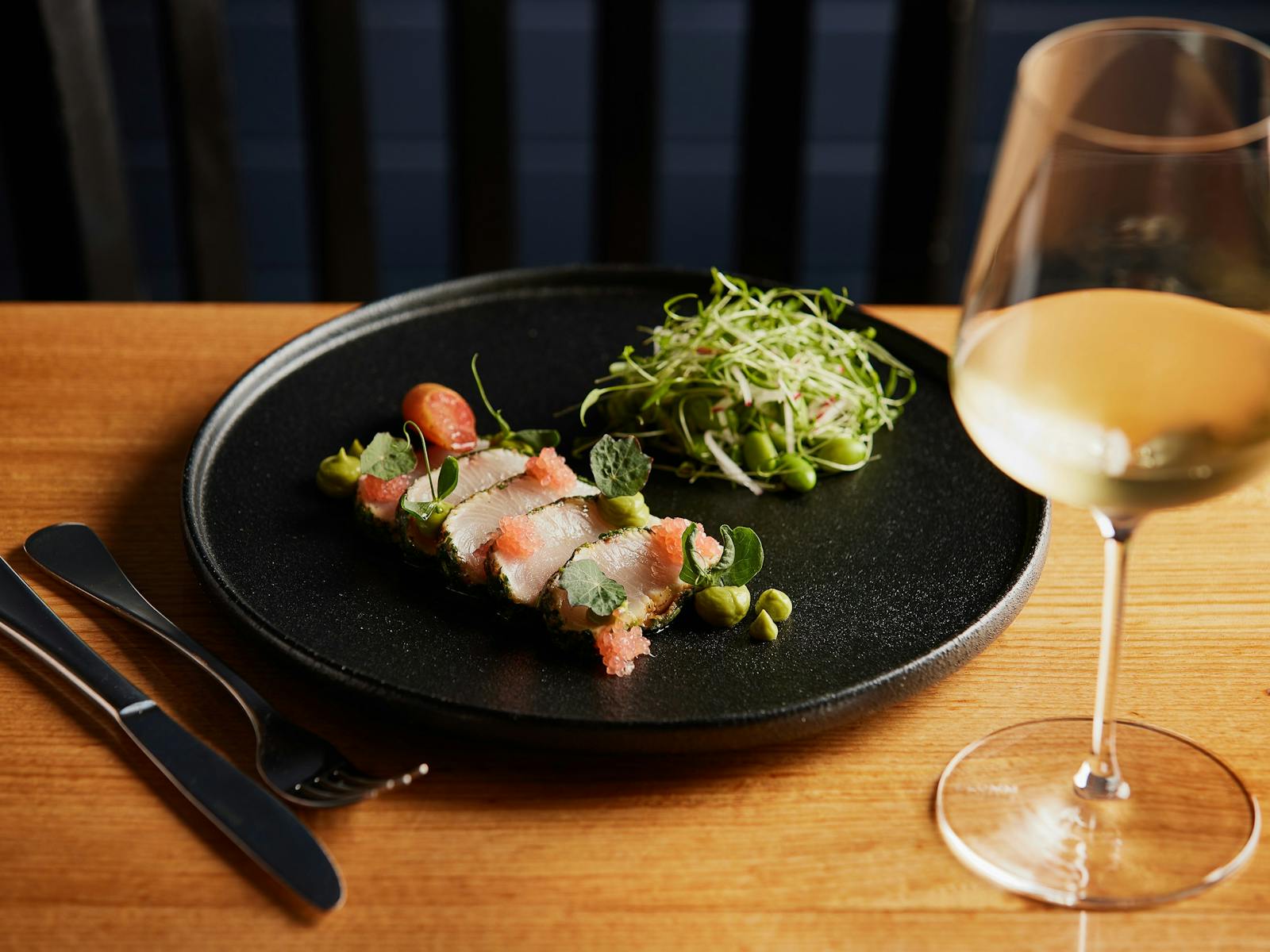 Australia Kingfish, thinly slice, presented on a black plate with a small salad and glass of wine.