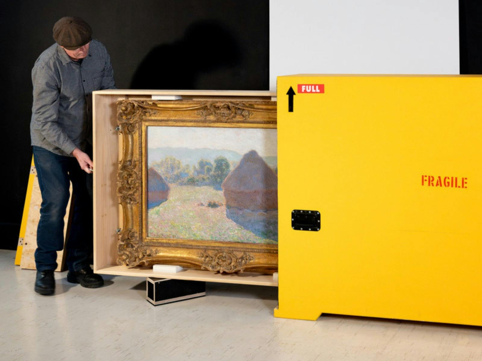 A man pulls a large painting of haystacks out of a a bright yellow crate marked fragile.