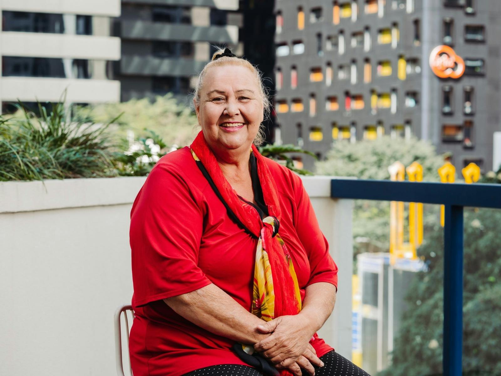 A smiling woman sits on a balcony, wearing a red shirt and a red and yellow scarf.