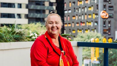 A smiling woman sits on a balcony, wearing a red shirt and a red and yellow scarf.
