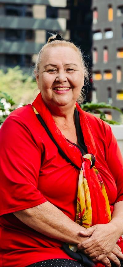 A smiling woman sits on a balcony, wearing a red shirt and a red and yellow scarf.