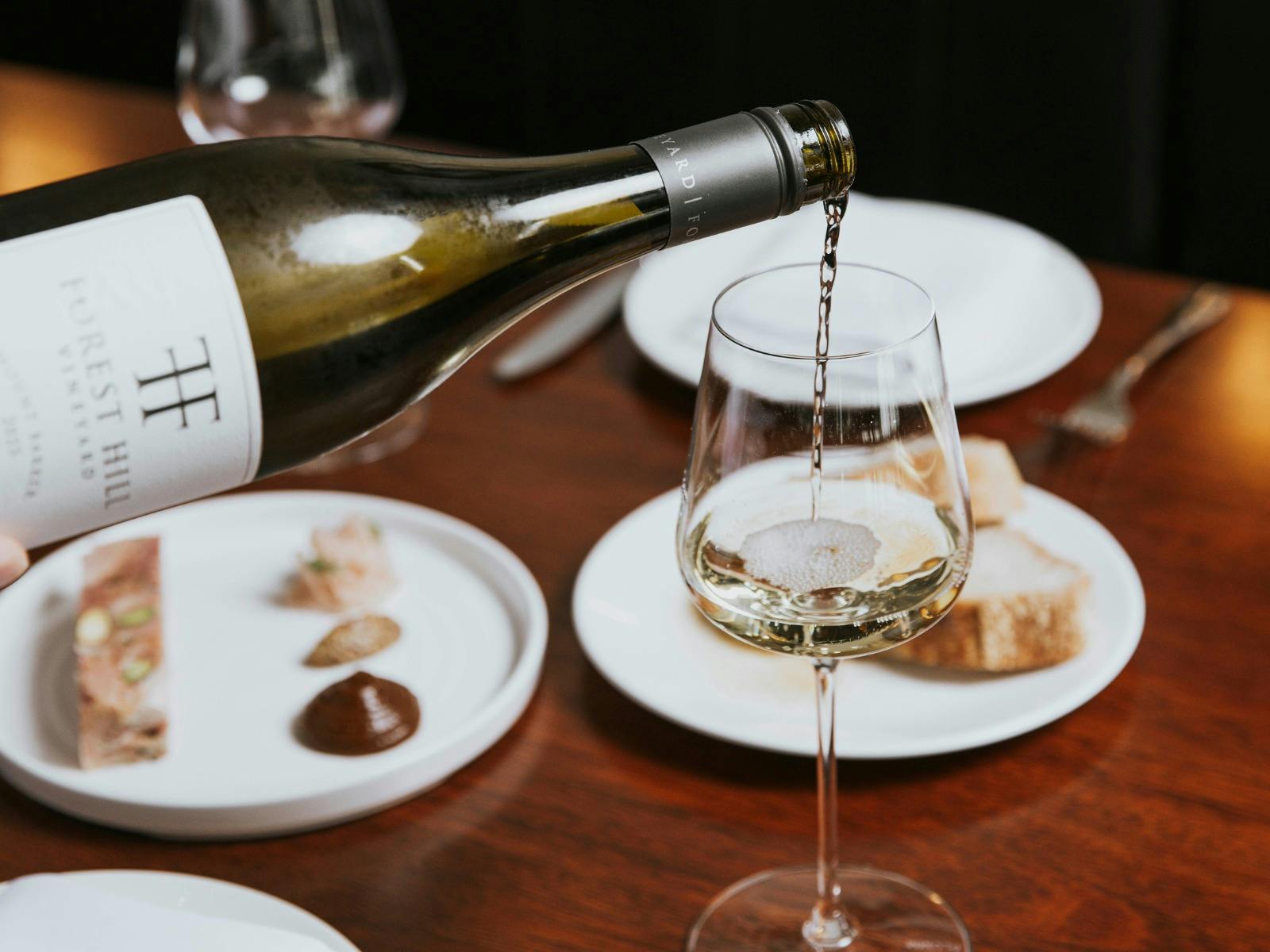 A glass of white wine being poured at the table, with plates of food out of focus in the background