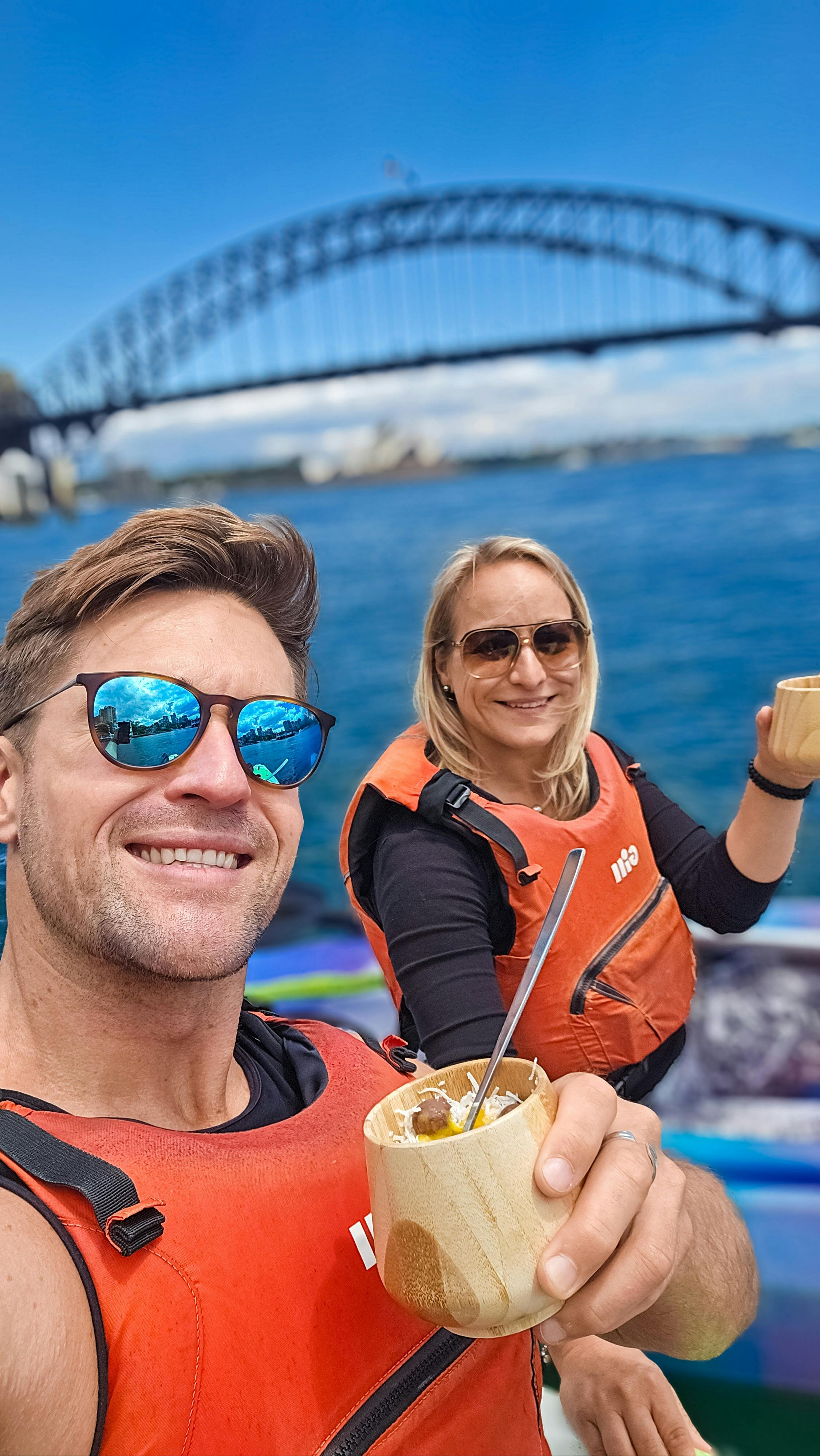 Man with blue sunglasses and lady with blonde hair sitting in kayaks eating ice cream opera house