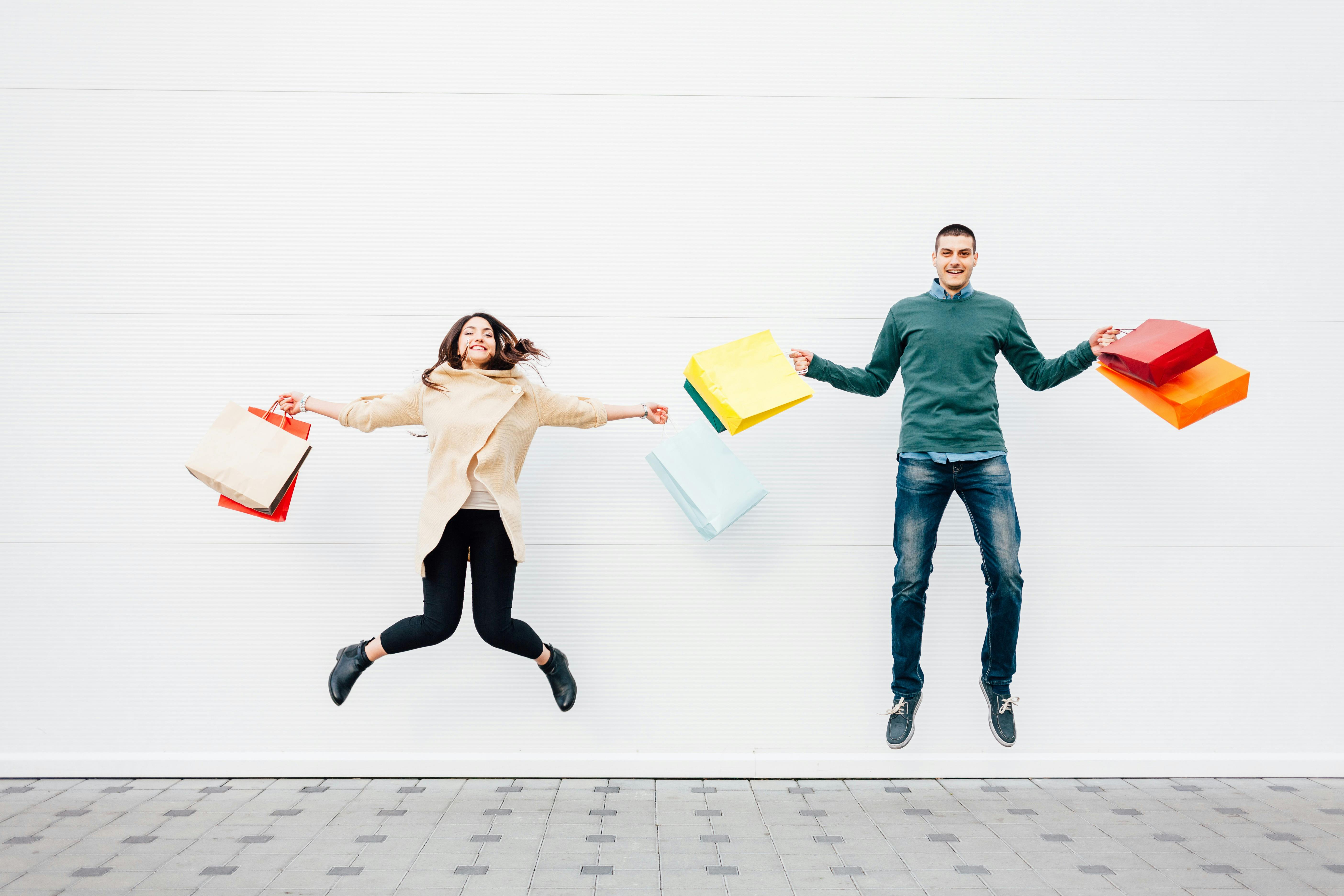 A man and a women holding shopping bags jumping in the air and smiling.
