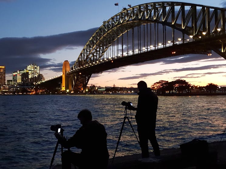 Setting sun on Sydney harbour with photographers in foreground looking towards the Harbour Bridge