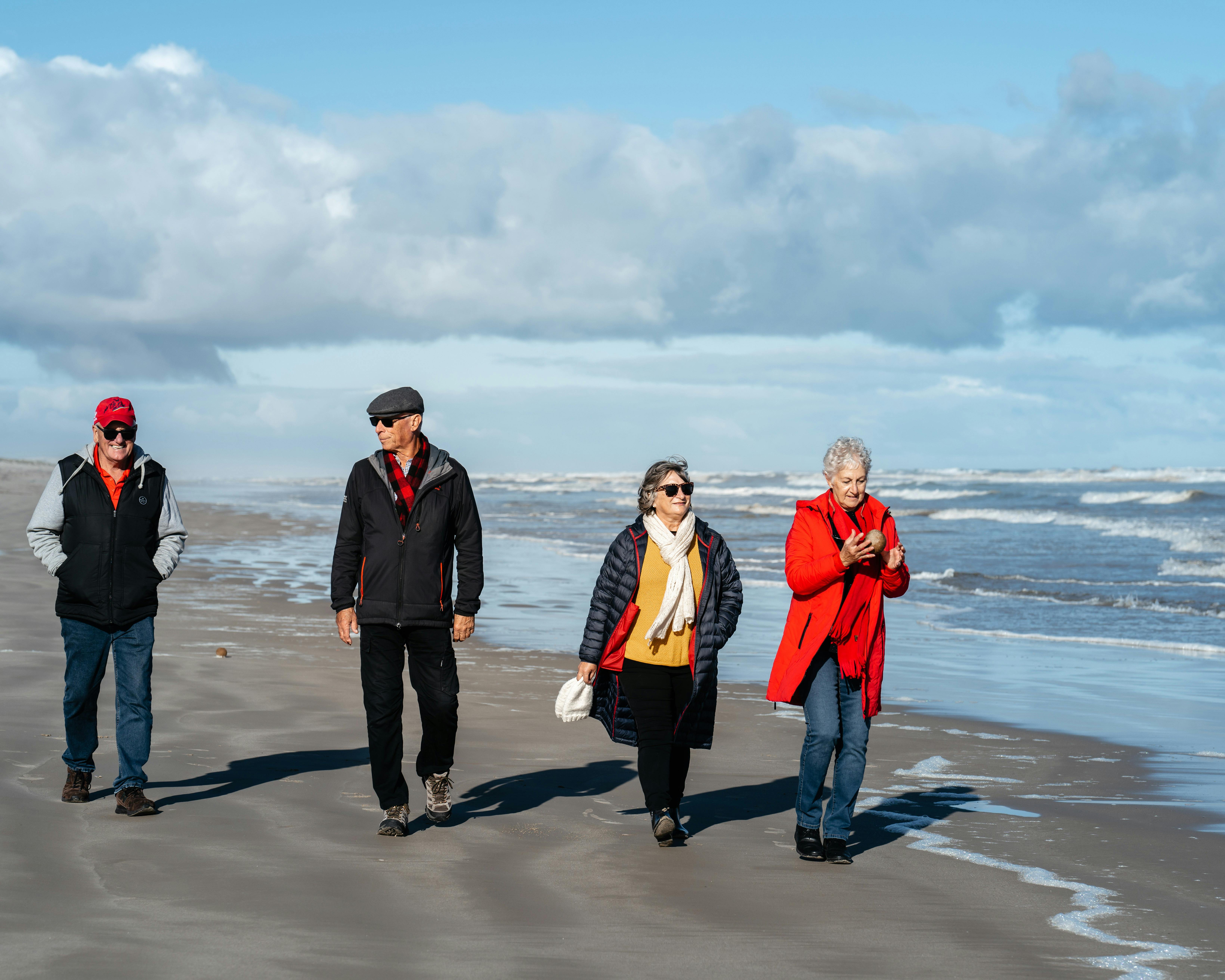 Four adults in jackets walk along a sandy beach by the ocean, enjoying a sunny, windy day.