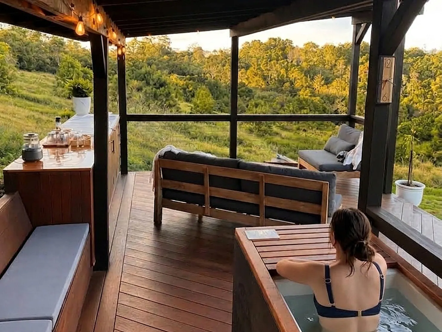 Woman in cold plunge on covered timber deck overlooking green hinterland valley at sunset