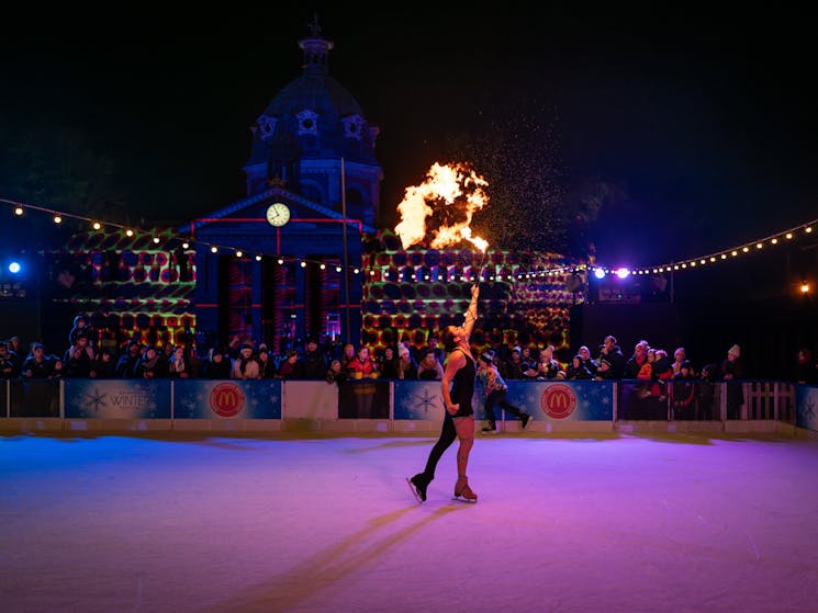 Ice skating performer blowing fire in front of crowds at the ice rink.