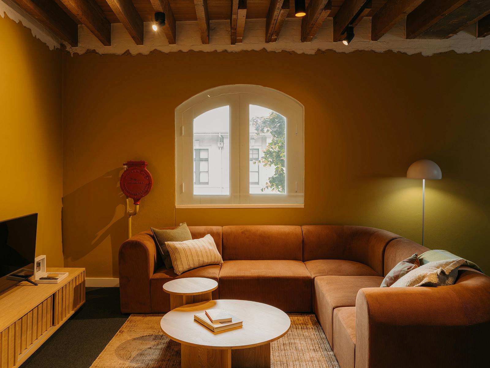 Cozy lounge with brown corduroy sofa, arched window, and warm mustard-toned walls.