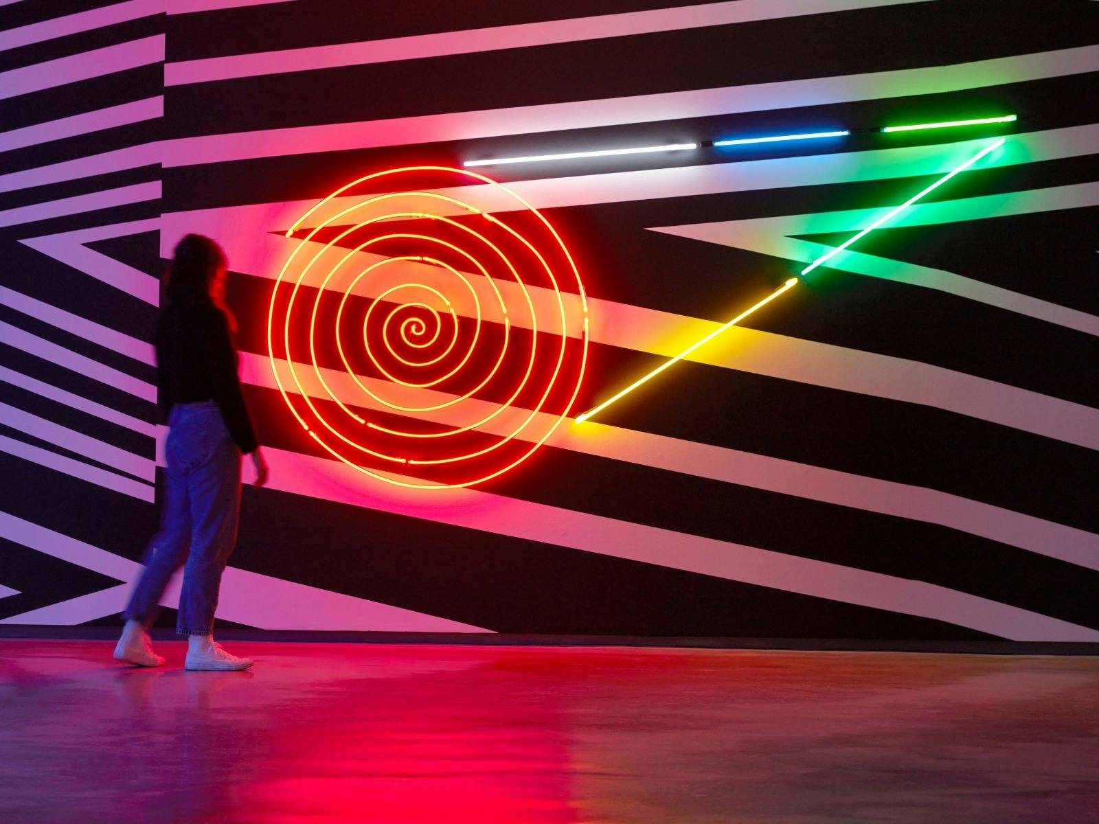 A person viewing an artwork installation  with graphic paint on a wall and a neon light overlayed.