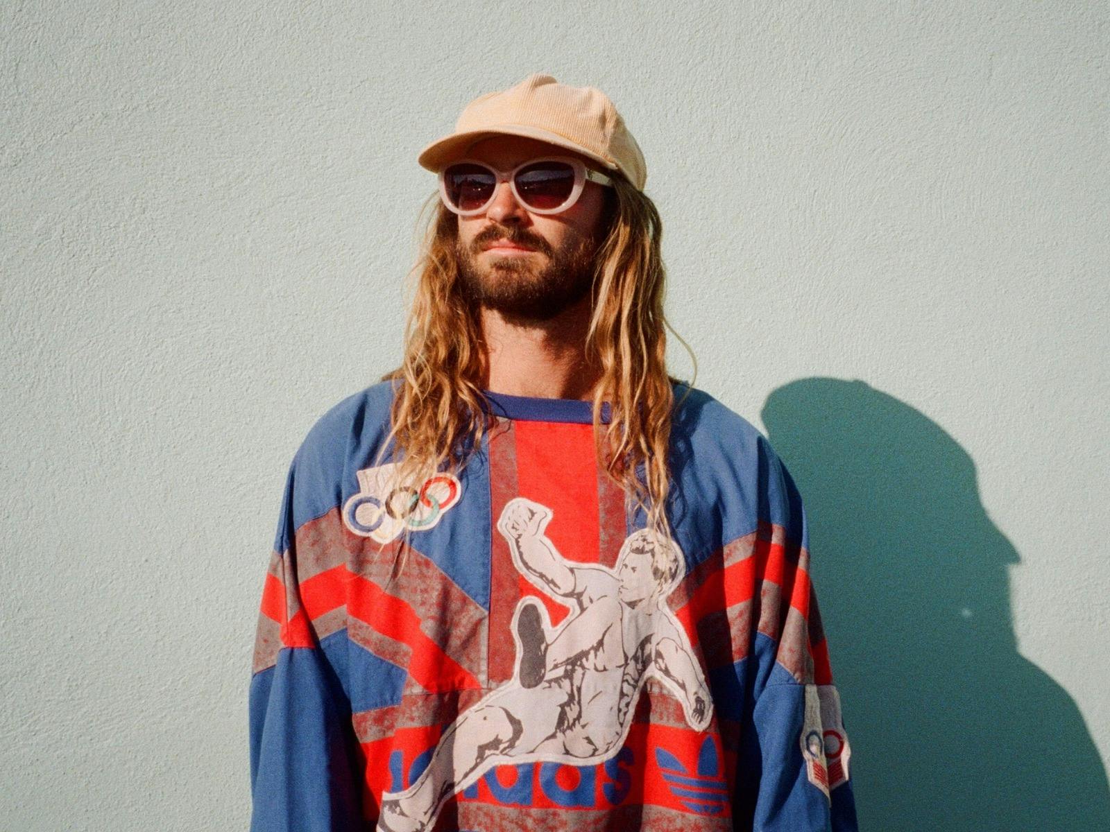A man with long, wavy hair and a beard stands in front of a pale blue wall, casting a shadow.