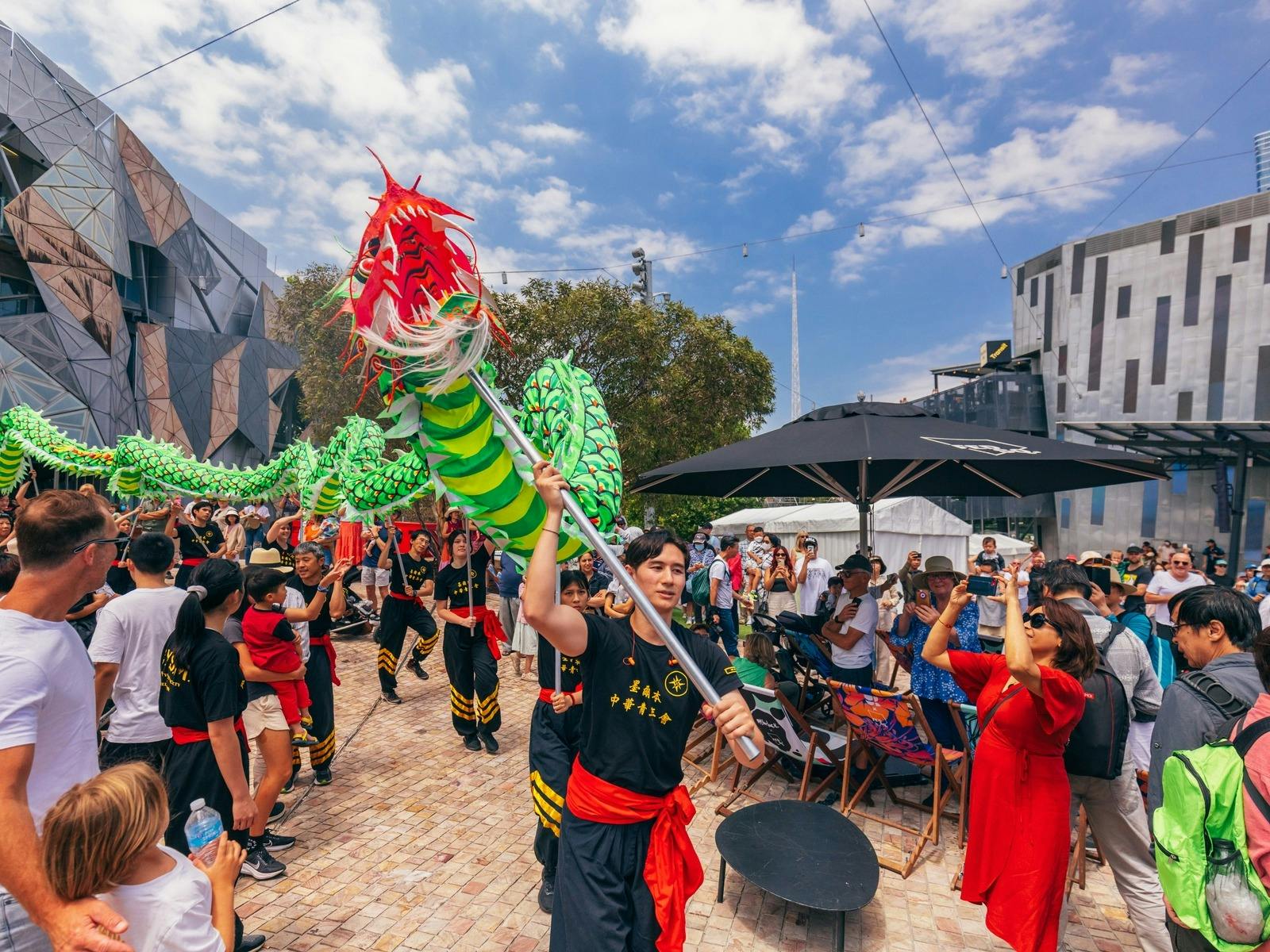 A group of young people move a green Chinese dragon through Fed Square, on a sunny day.