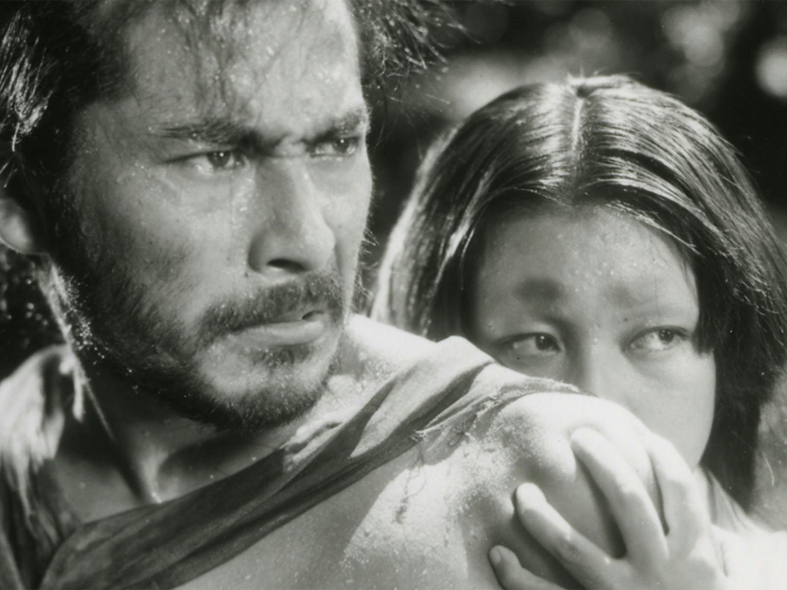 Black and white still of a man looking angry and a woman hiding behind the man