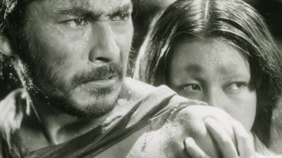 Black and white still of a man looking angry and a woman hiding behind the man