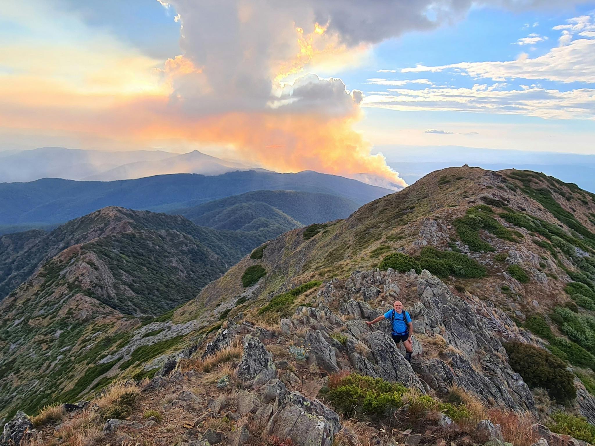 A hiker heading up West Ridge of Mt Buller with smoke rising towards the sky in the background.