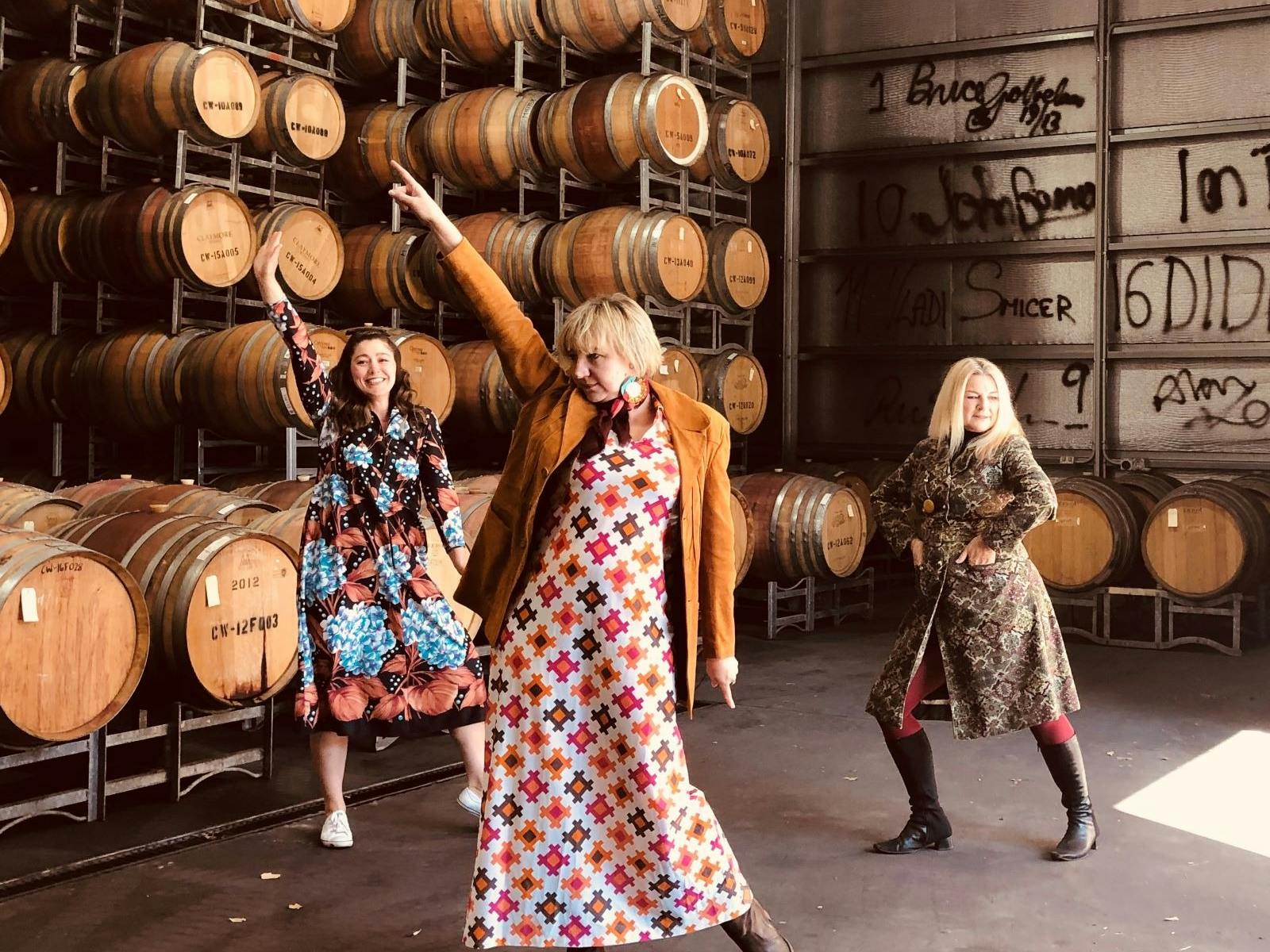 three women in 1970's dresses dance among barrels of wine